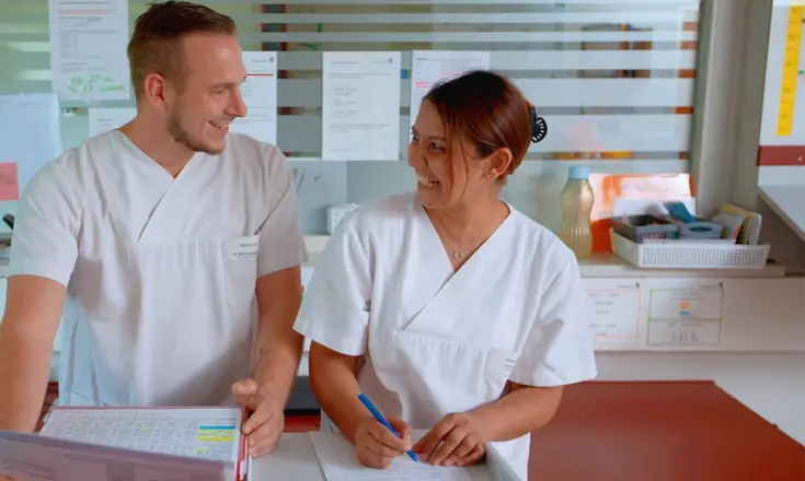Neslihan Cali steht mit einem Kollegen an einem Tisch, auf dem Arbeitsblätter und -ordner liegen. Beide tragen weiße Kittel und lachen sich an.