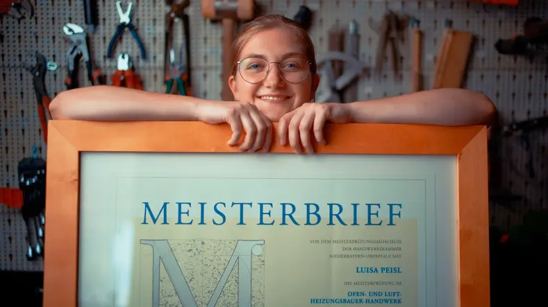 Luisa Peisl lächelt und stützt ihre Arme auf ihren gerahmten Meisterbrief, der vor ihr auf dem Tisch liegt.