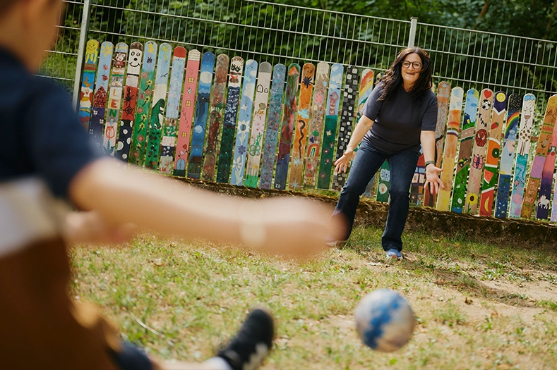 Lilli Fischer steht mit ausgebreiteten Armen vor einem bunten Zaun, während ein Junge ihr einen Ball zuspielt.