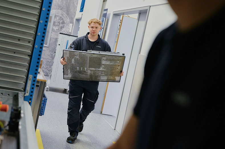 Ein jünger Mann in Arbeitskleidung trägt ein Maschinenteil in den Händen.