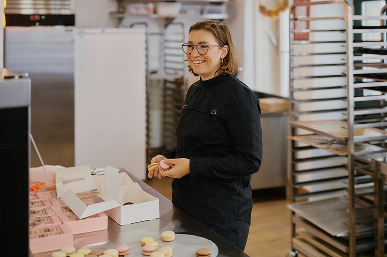 Laura Schönberger trägt eine schwarze Kochuniform und hält lachend zwei Macarons in den Händen.