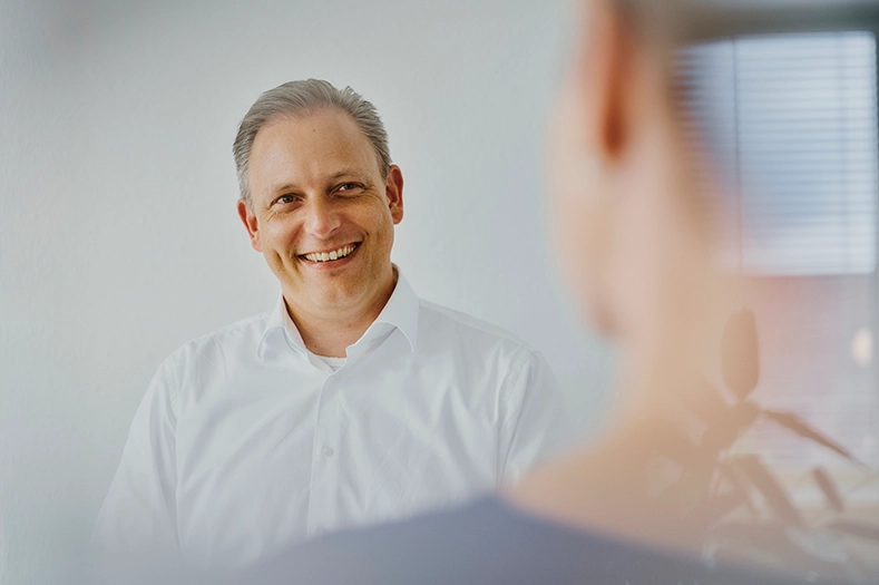 Dirk Egelseer trägt ein weißes Hemd und lächelt freundlich in die Kamera. Er ist im Gespräch mit Tina Leitschuh. 