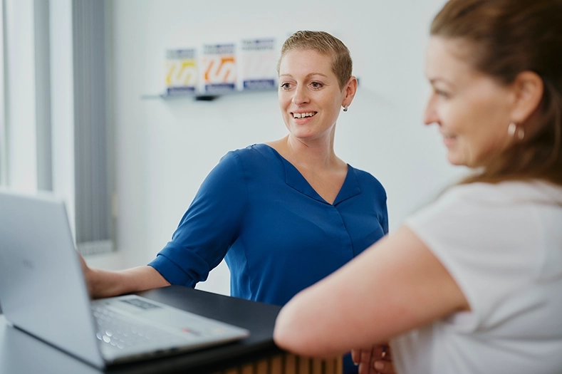 Tina Leitschuh trägt eine königsblaue Bluse. Sie steht vor einem Laptop und unterhält sich mit einer Frau. 