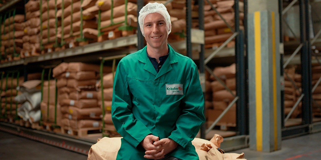 Michael Herrmann trägt einen grünen Kittel und eine Schutzhaube. Er sitzt in einer Lagerhalle vor gestapelten Säcken.