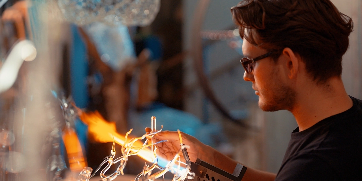 Michael Weinfurtner trägt eine Brille und bearbeitet mit einem Werkzeug ein Stück glühendes, heißes Glas.