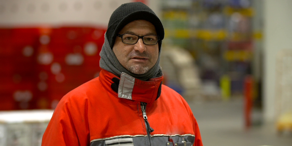 Roland Jahn trägt eine rote Arbeitsjacke und eine Mütze. Er steht in einem Lager und blickt in die Kamera.