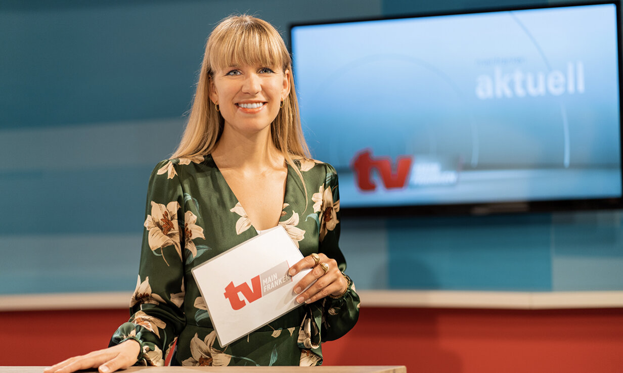 Sandra Hechelmann vor großem Display stehend, hält Moderationskarten mit der Aufschrift TV in der Hand.