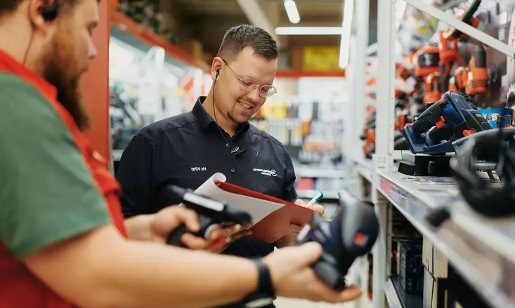 Bernhard Kittl hält einen Kugelschreiber in der Hand und macht Notizen auf einem Klemmbrett. Links sieht man einen Mann mit einem kleinen Gerät in der Hand.