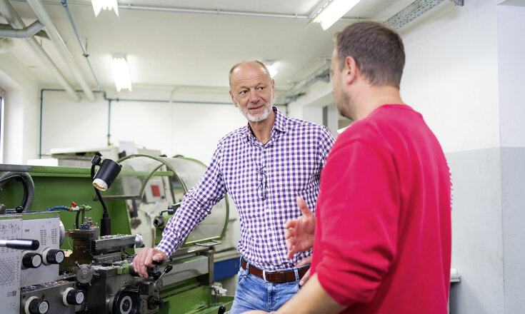 Ralf Greif und ein Mitarbeiter stehen in einer Werkstatt und unterhalten sich.