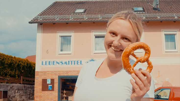 Frau Schönberger steht lächelnd vor ihrer Bäckerei und hält eine Breze in die Kamera.