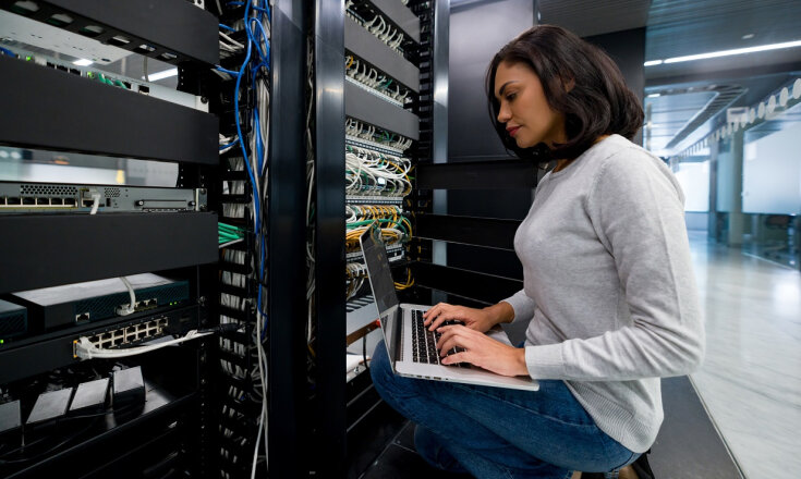 Eine Frau sitzt mit einem Laptop auf dem Schoß in einem Raum mit IT-Equipment.