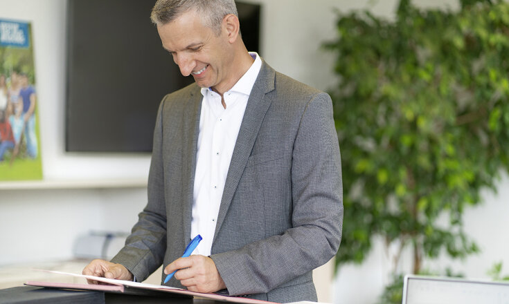 Werner Müller schaut lächelnd auf ein Dokument und hält einen Stift in der Hand."
