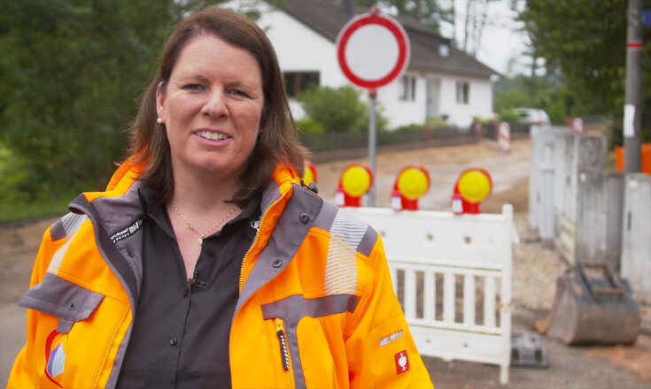 Kerstin Ludwig trägt eine orangefarbene Sicherheitsjacke und steht lächelnd auf der Straße vor einem Bauzaun.